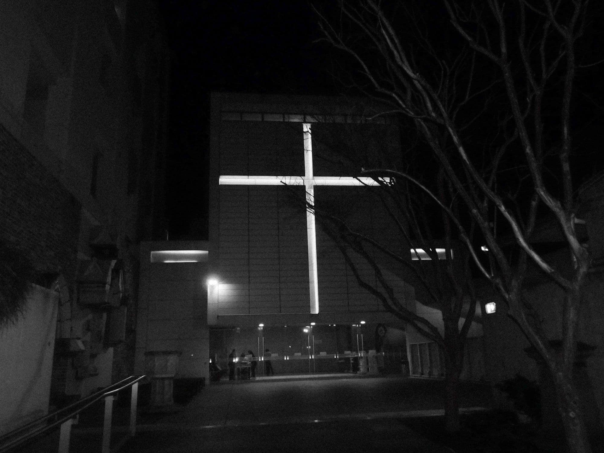 Night photos of St Barnabas on Broadway, its modern facade marked by a huge illuminated cross, seen through bare tree branches, from the forecourt, and looking up at the building.