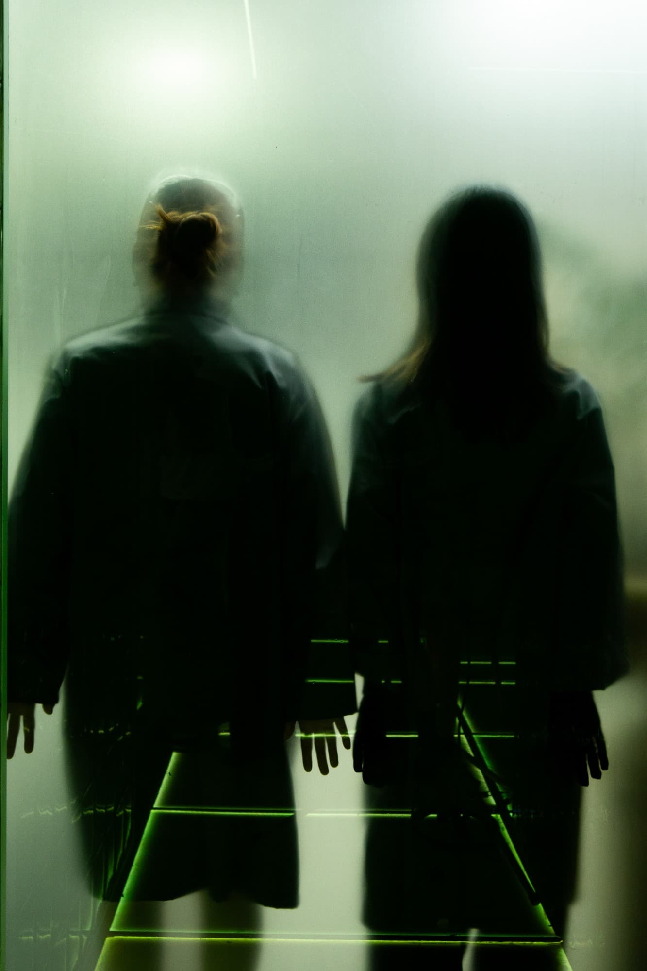 Two blurred figures seen from behind through frosted glass, above green-lit steps.