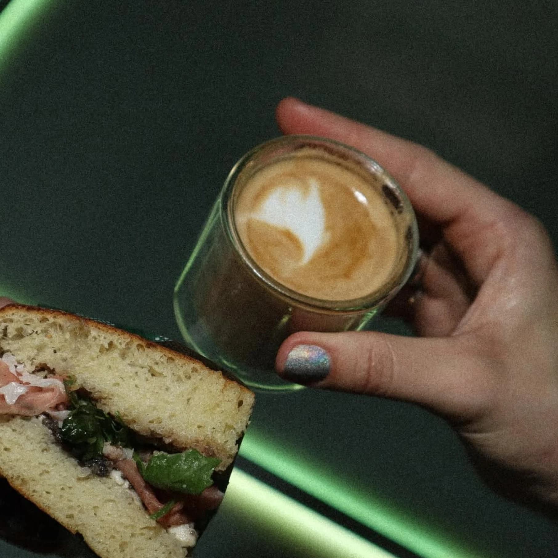 A hand holding a glass of latte beside a focaccia sandwich under green light, then a black card reading "If we go dark, tell the cops it was the focaccia".